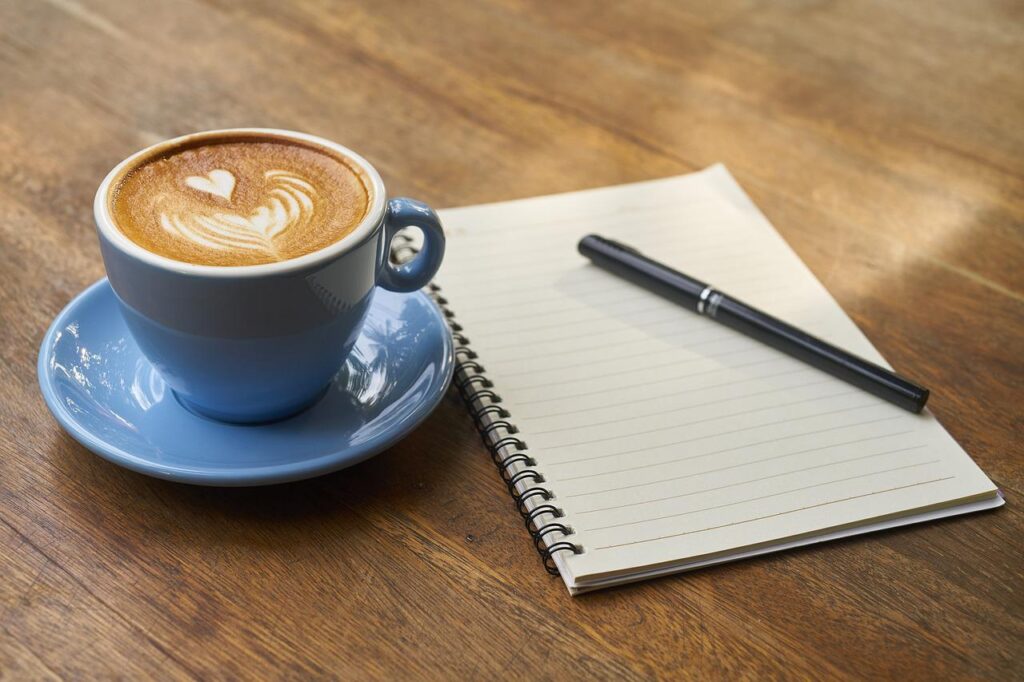 A cup of cappuccino in a blue mug placed on a blue saucer with a spiral notebook and a black ballpen on a brown wooden table