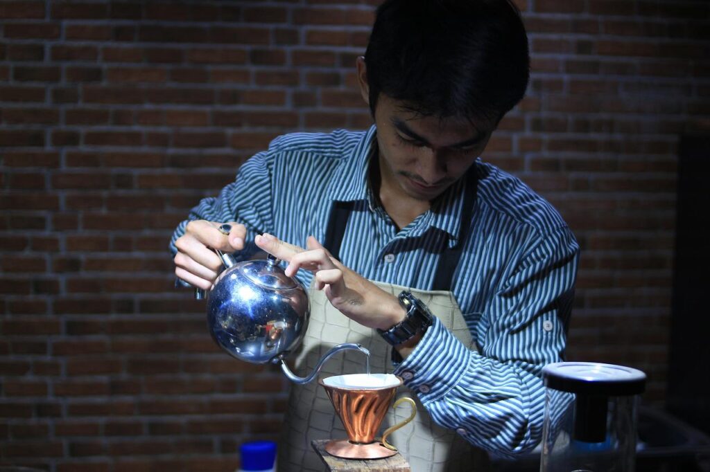 Hot water in a stainless kettle is being poured to make coffee by a young man wearing a striped blue and white long sleeves shirt