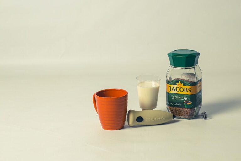Jacobs' instant coffee and a milk frother beside a cup of milk and a brown mug on a white surface