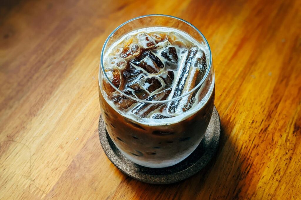 An iced coffee in a glass placed on a black ceramic coaster on a wooden table