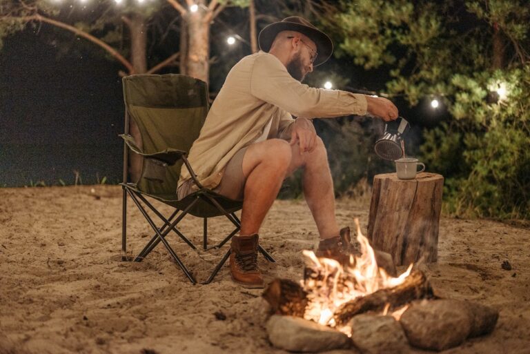 A man wearing a brown hat and brown long sleeves sitting in a brown camping chair near a bonfire in a forest