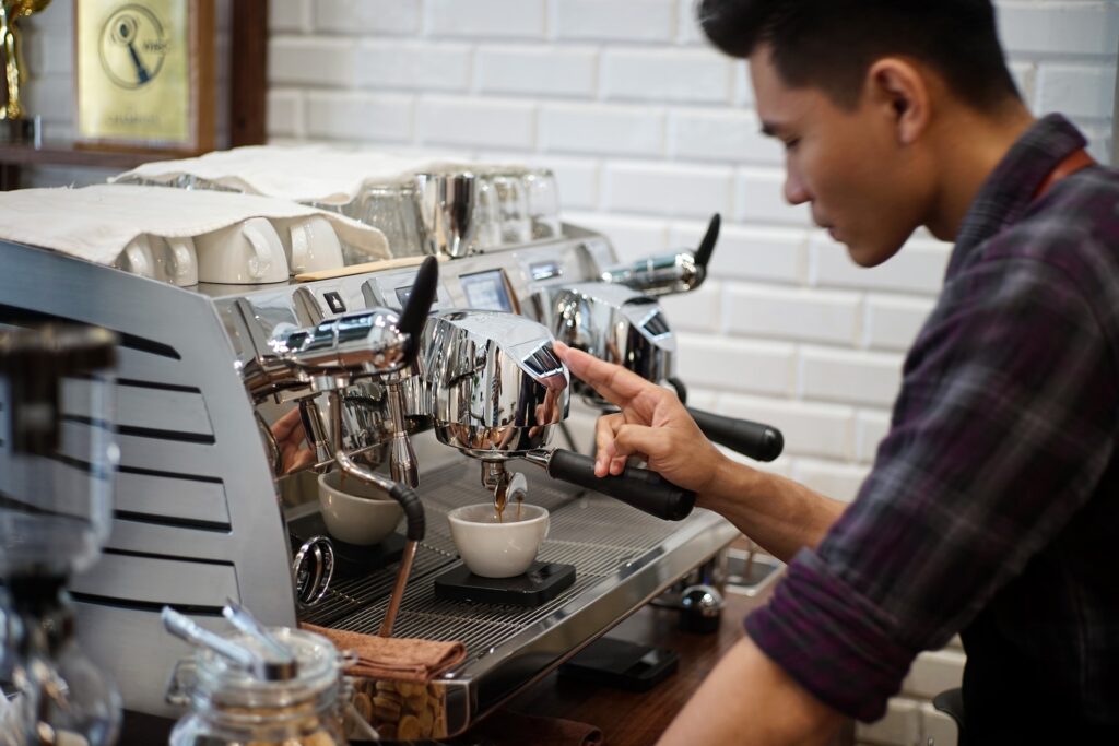 A young barista making a fresh cup of coffee using a silver coffee maker inside a cafe 