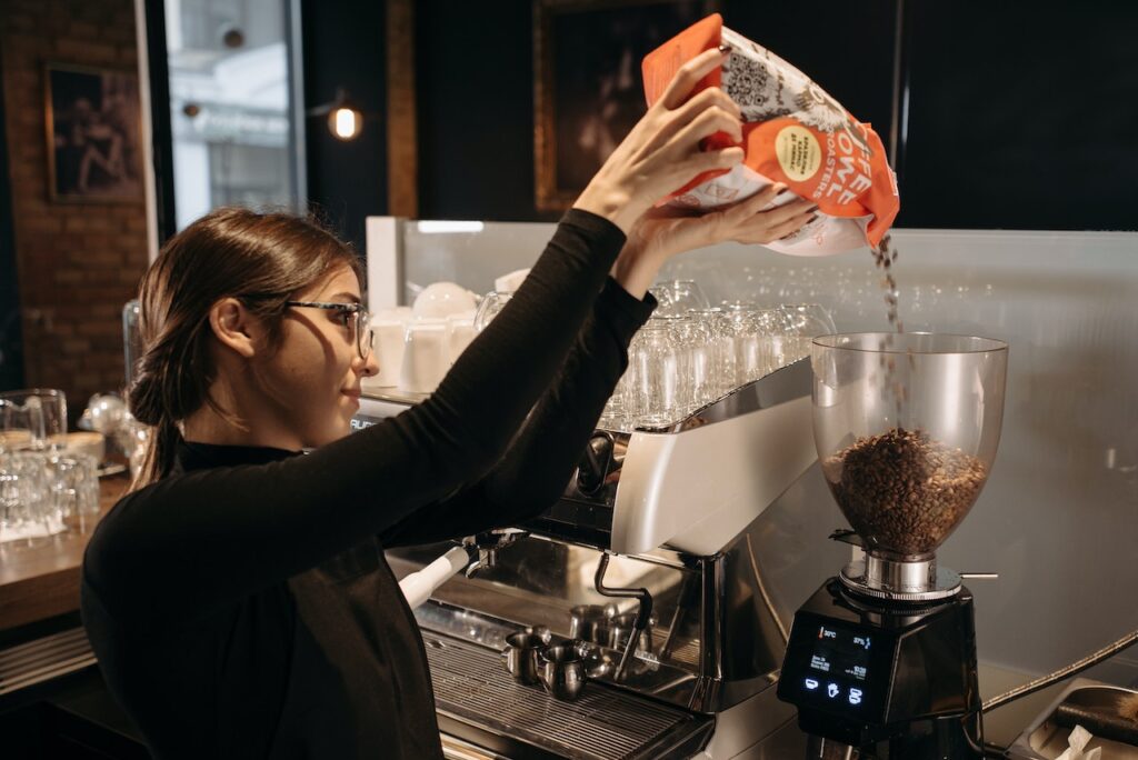 A woman wearing black long sleeves and eyeglasses puts whole coffee beans on a black coffee grinder in a coffee shop