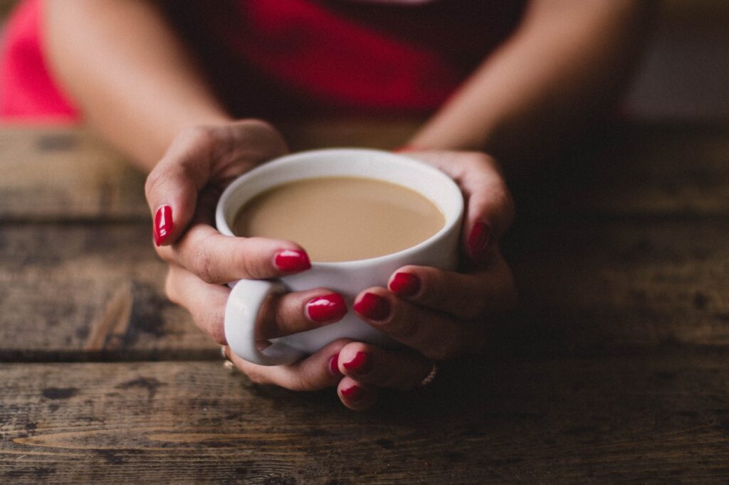 A person with red nail polish holds a white coffee mug placed on a brown wooden table