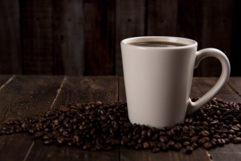 A white mug filled with black coffee surrounded by coffee beans on a brown wooden table