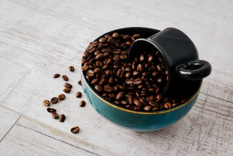 A black mug scooping whole coffee beans placed in a blue bowl on a white wooden table